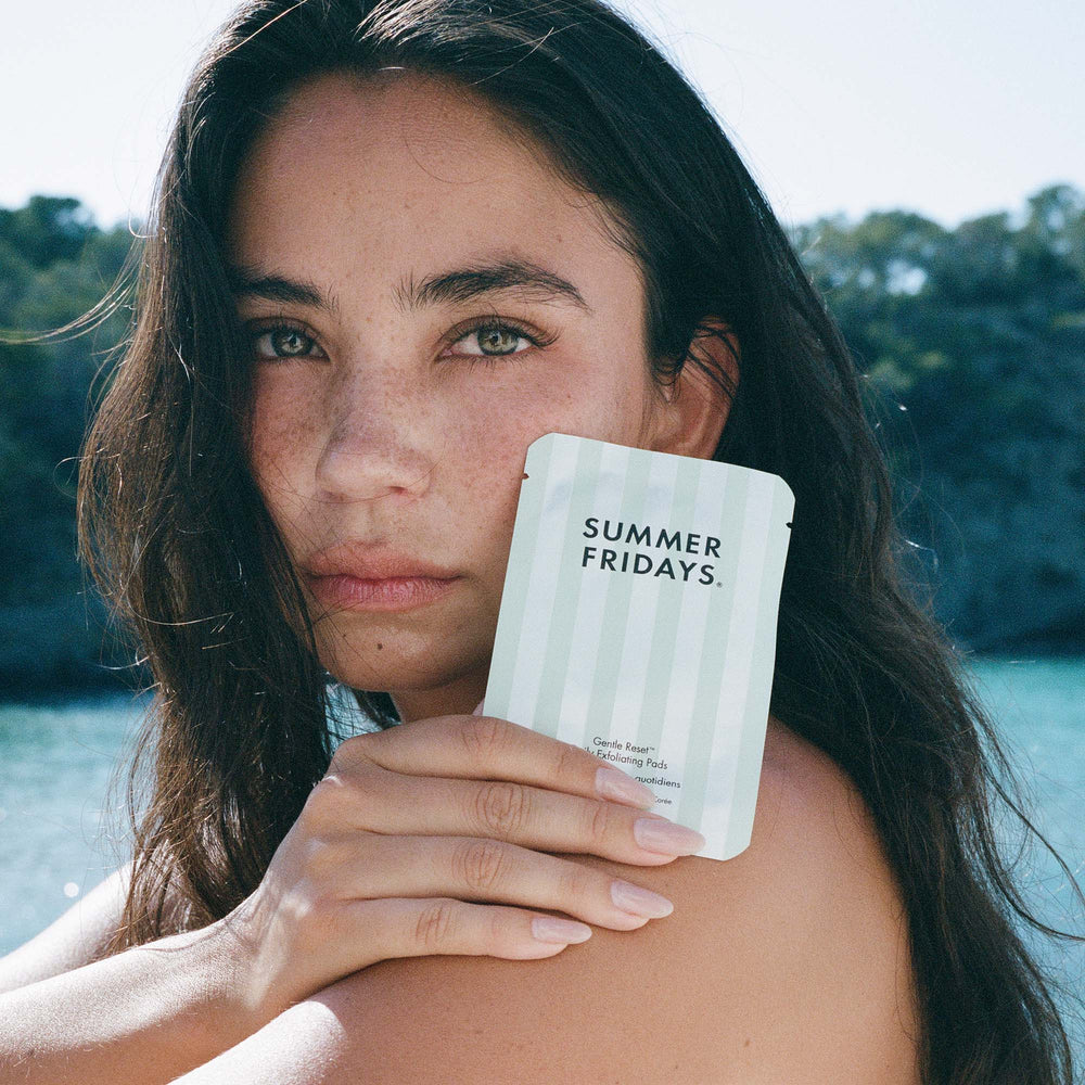 Woman holding a 'Summer Fridays' product outdoors with water and greenery in the background