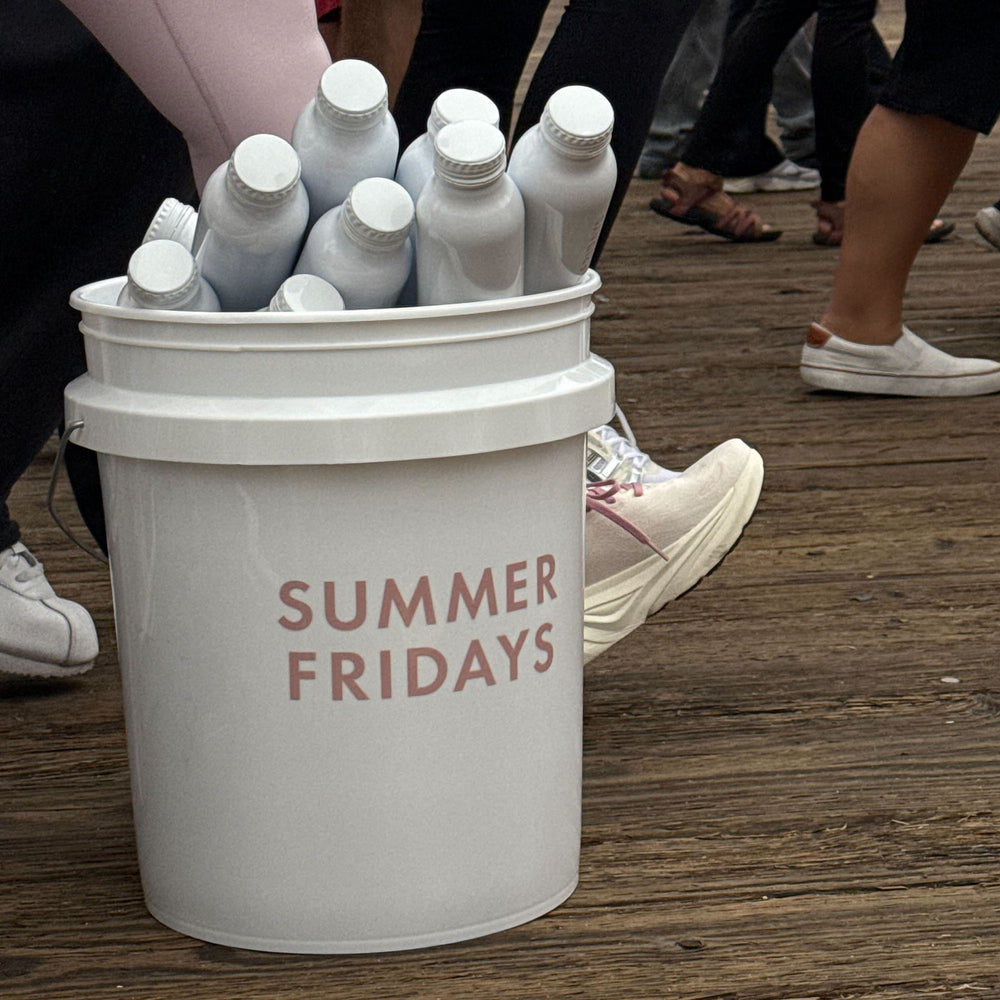 White bucket labeled 'Summer Fridays' filled with white bottles on a wooden floor.